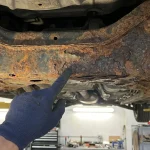 Une main gantée pointe une zone de rouille perforante et de corrosion avancée sur le châssis métallique d'un véhicule.