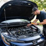 Un mécanicien utilise un multimètre pour tester la batterie d'une voiture bleue avec le capot ouvert dans une allée.