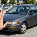Une main tendue faisant un signe d'arrêt devant une berline grise garée dans une rue résidentielle.