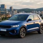 Un SUV Volkswagen T-Roc bleu est garé sur une route sinueuse avec un panorama urbain de gratte-ciel au crépuscule.