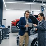Un inspecteur en veste et une mécanicienne en combinaison examinant une tablette devant une voiture dans un garage.