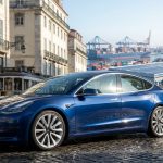 Une Tesla bleue garée dans une rue pavée européenne avec un port maritime et des porte-conteneurs en arrière-plan.
